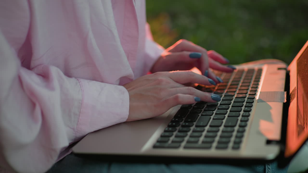 primer plano de una mujer con una camiseta rosa con uñas bien pulidas escribiendo en una computadora portátil, sombras de su mano visibles en el teclado, con un cálido resplandor de luz solar en el fondo, efecto de luz bokeh en la distancia