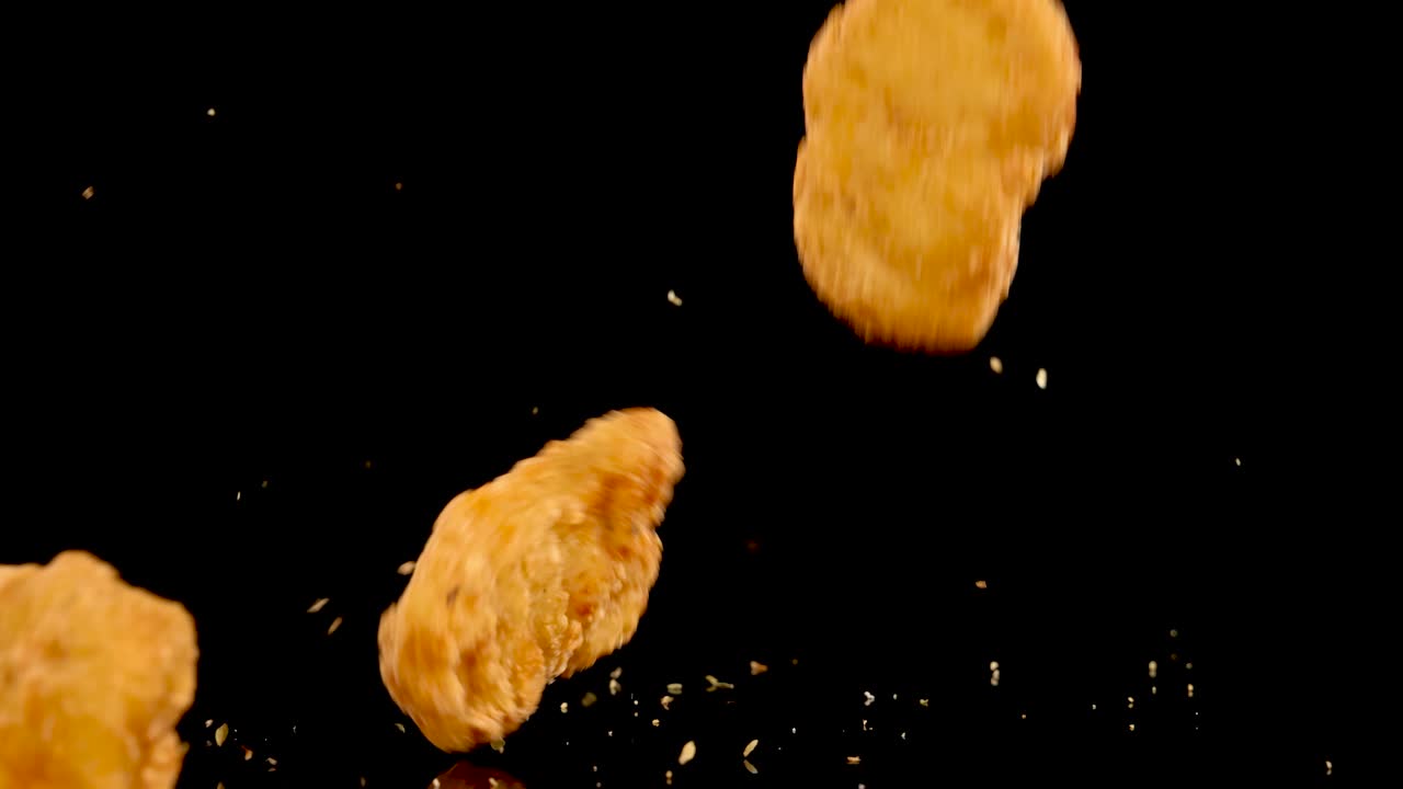 Golden breaded chicken nuggets tumble and spin against a black background, scattering crumbs midair. Dramatic lighting and high-speed camera capture crisp texture and dynamic motion