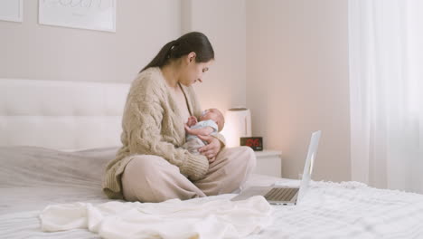 棕色女人坐在笔记本电脑前面的床上,抱着她的婴儿并亲吻他