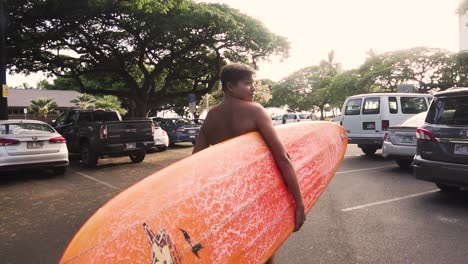 young boy walks through parking lot holding red longboard