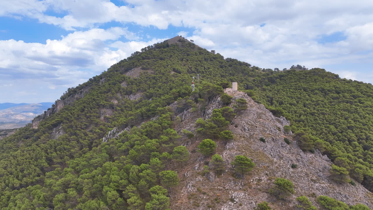 Aerial flies towards cell tower and ancient watch tower atop green forested hill