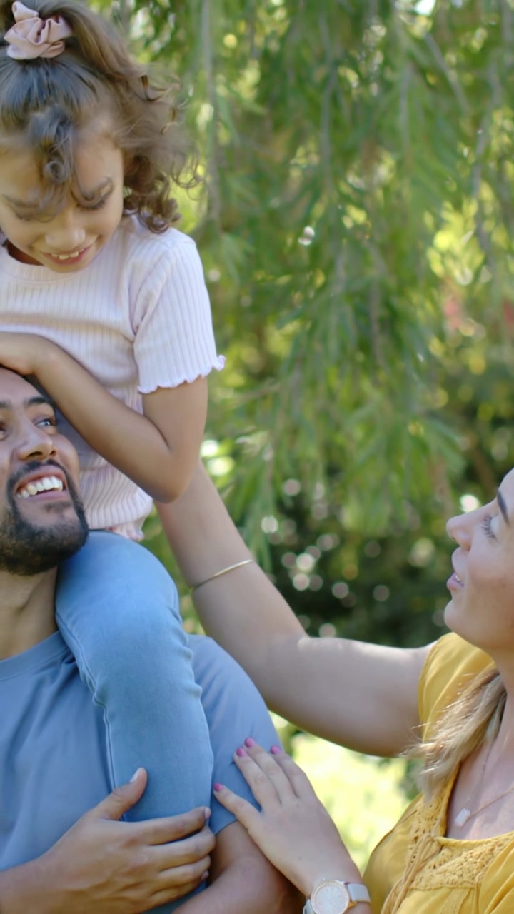 Vídeo vertical: Pai brincando carregando a menina nos ombros no parque enquanto a menina coloca as mãos na cabeça, observando.