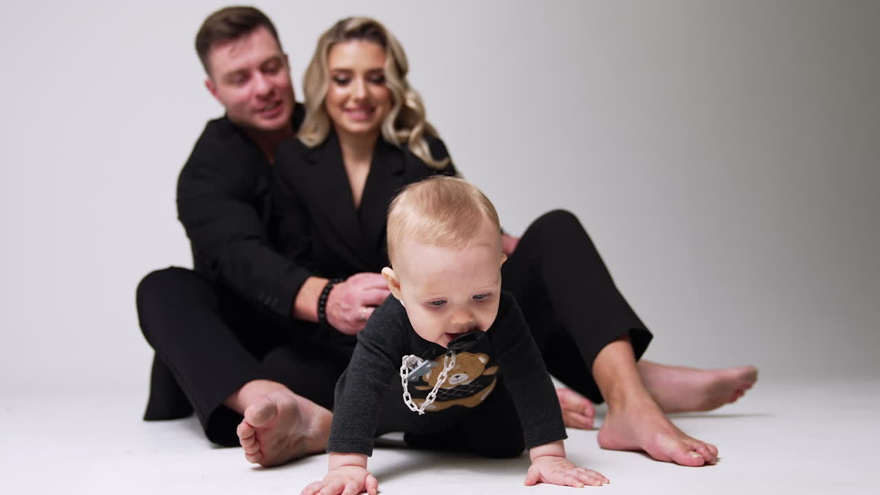 Mom and dad wearing black suits sit on the floor in studio. Parents look at their adorable baby trying to crawl up to camera. Blurred backdrop. White backdrop.