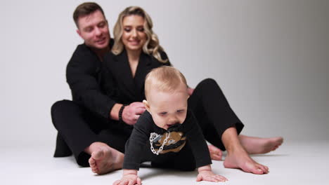 mom and dad wearing black suits sit on the floor in studio. parents look at their adorable baby trying to crawl up to camera. blurred backdrop. white backdrop.