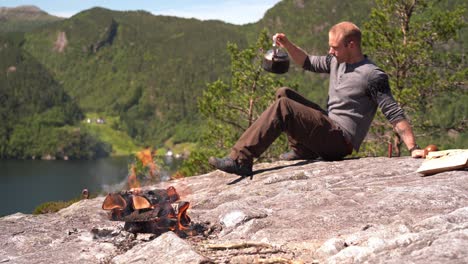 man sitting down on rock an pouring himself a cup of freshly brewed coffee with mountain scenery in background and bonfire in foreground - european male in lush summer mountain nature