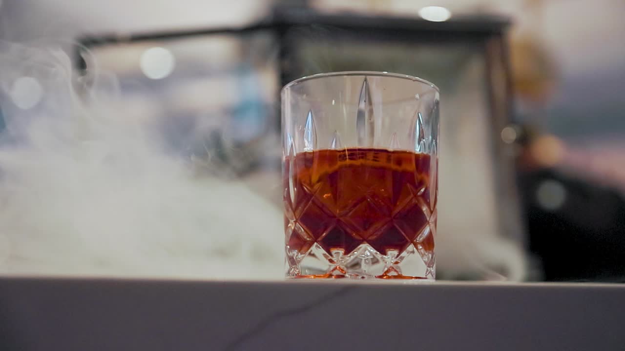 Slow motion video of a smoking whiskey drink resting on a bar. Smoke swirls gently around the glass, creating a cinematic and moody atmosphere