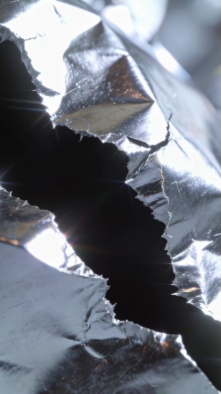 Vertical video: Light shifting revealing jagged foil hole in close-up macro, showing frayed edges