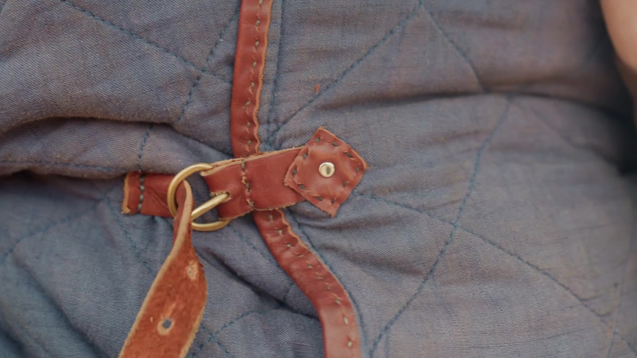 Close-up low-angle view shows hands threading brown leather belt through brass buckle on quilted blue-gray garment, with fingers pulling strap tight over dark coat for secure fit, with belt, leather