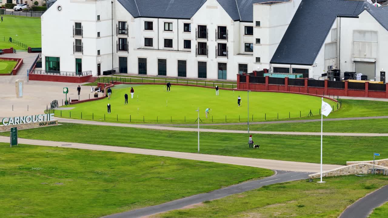 Golfers and a dog walker move across putting green near clubhouse, daylight, wide aerial view
