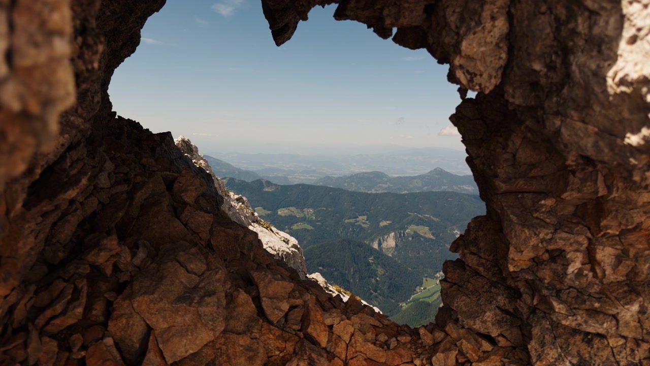 Blick durch eine natürliche Felsöffnung offenbart ferne Berge, bewaldete Täler und blauen Himmel, wodurch ein dramatischer Rahmen in einer hochalpinen Landschaft entsteht