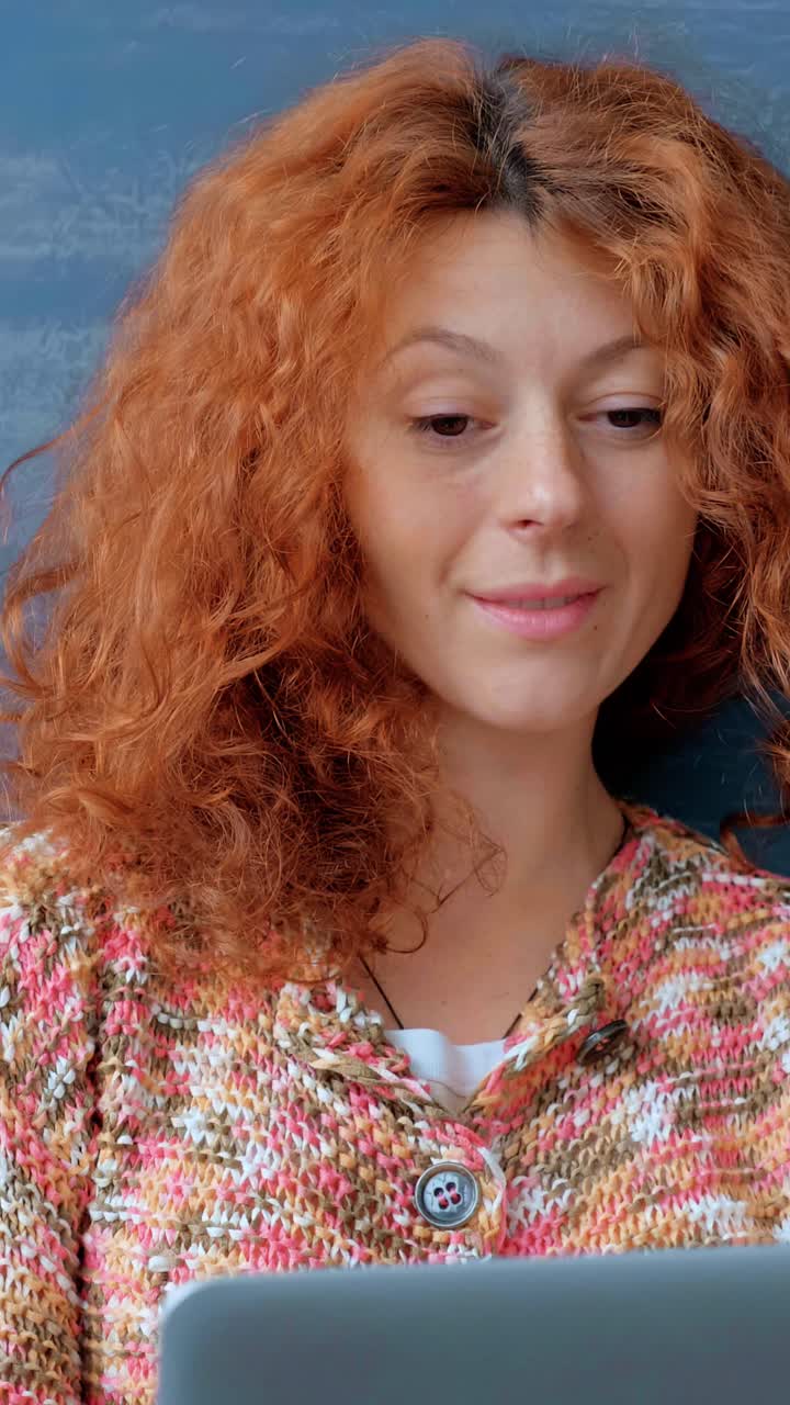 Woman with curly red hair is smiling while using laptop, wearing colorful knitted sweater, showcasing a relaxed and engaging work-from-home atmosphere