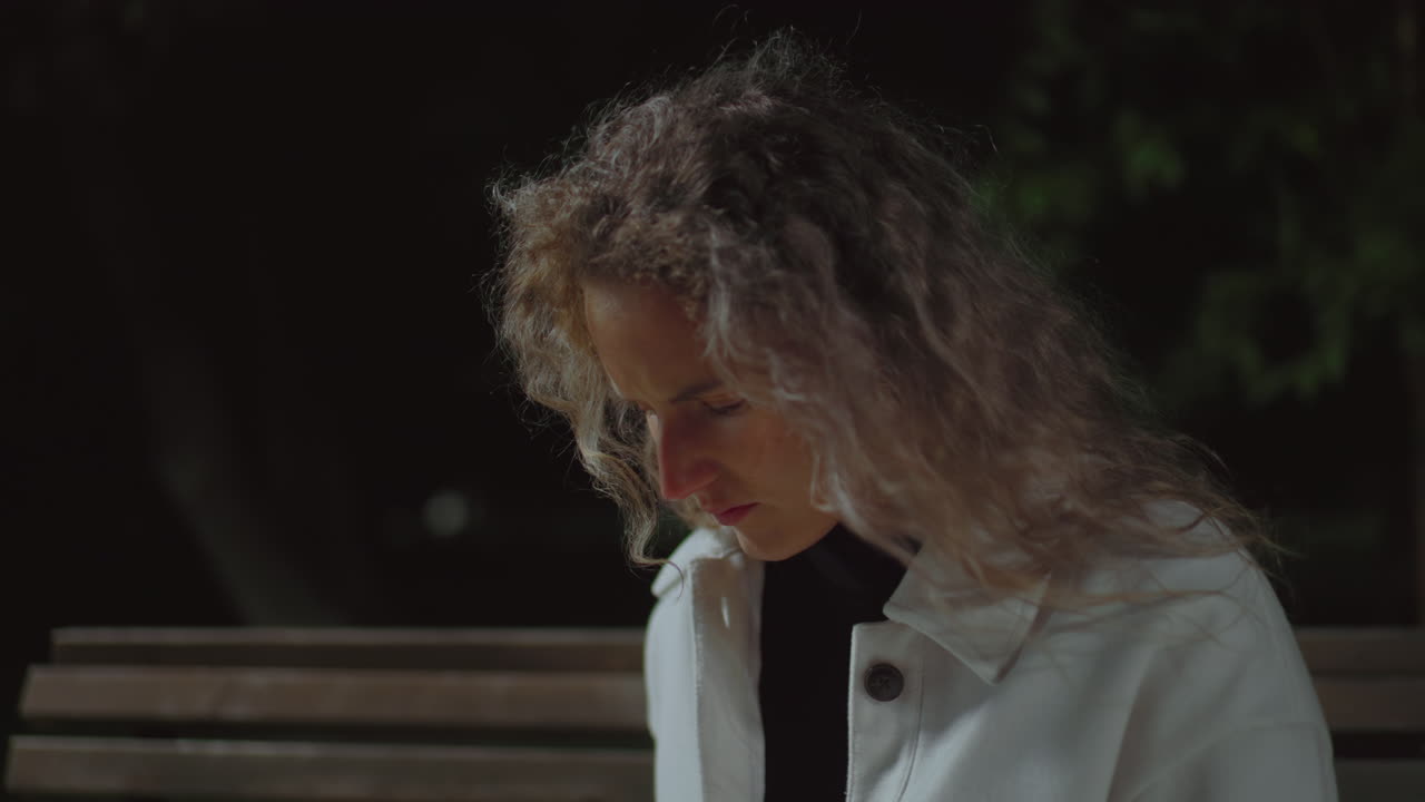 Young girl focuses on something thoughtfully, looks up with serious face wearing white coat and curly hair, seated on wooden bench at late evening under soft dim light in quiet outdoor park