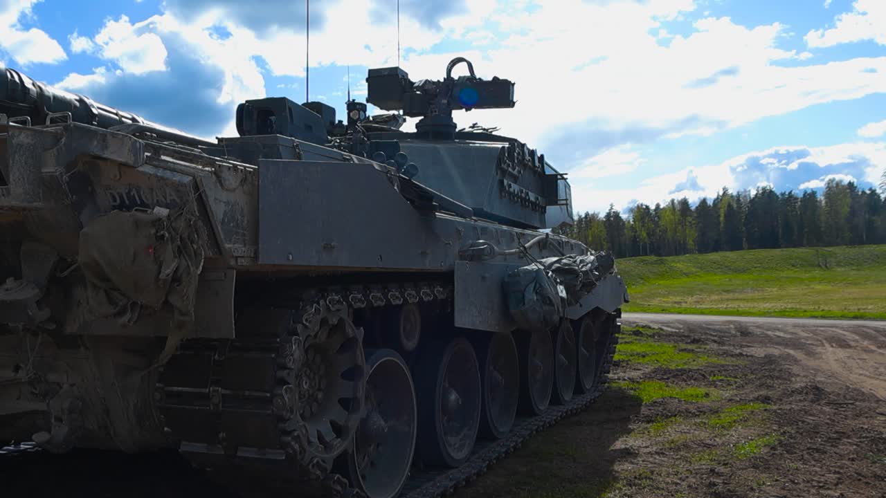 Close up or closeup footage of a British Challenger 2 4034 tank with its large cannon weapon and steel plates parked and standing on a grassy green field with mud during a sunny day, forest visible.