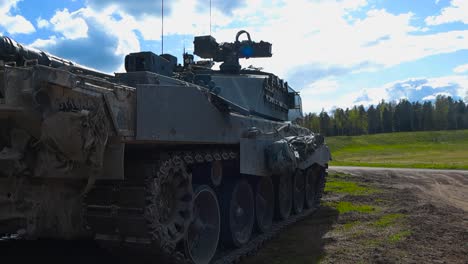 close up or closeup footage of a british challenger 2 4034 tank with its large cannon weapon and steel plates parked and standing on a grassy green field with mud during a sunny day, forest visible.