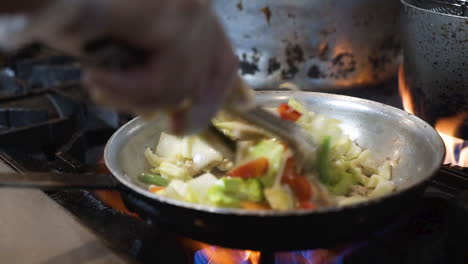 chef uses tongs to stir fry vegetables in pan over high heat flames in commercial restaurant kitchen, close up hd