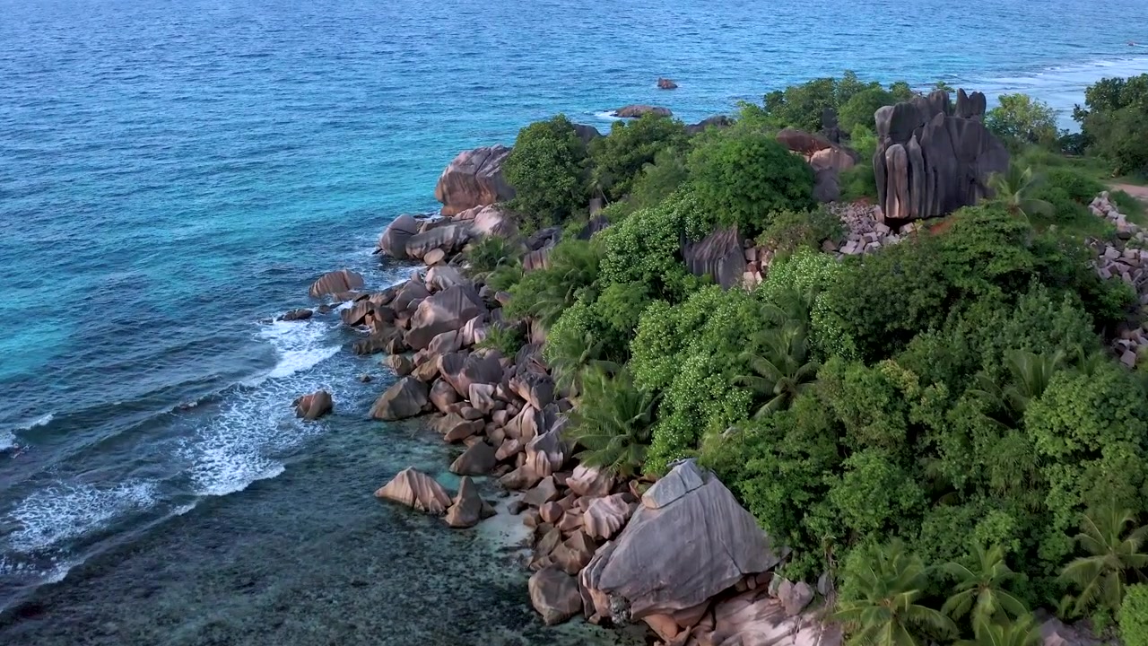 Ripresa aerea con drone in panning lungo una spettacolare costa delle Seychelles con iconici massi di granito, alberi tropicali, una capanna con tetto di paglia, mare turchese e spiaggia di sabbia bianca in primo piano