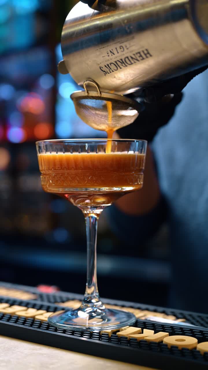 New York, USA, 15 aprile 2026: Barman che versa un cocktail all'arancia attraverso un colino in un bicchiere. Liquido che scorre dallo shaker attraverso un colino a maglie in un bicchiere coupe di cristallo su un bancone da bar.