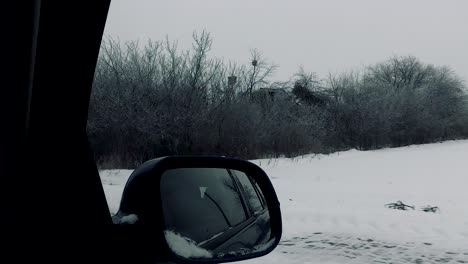 Pov-of-car-side-mirror-travel-passenger-looking-at-roadside-landscape-through-dirty-window-winter-scenery-snow-forest-bare-trees-cloudy-sky