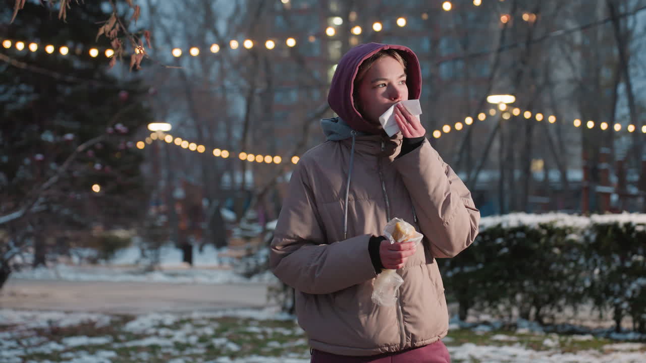 Una donna che tiene uno spuntino in mano e usa un tovagliolo per pulirsi la bocca in un ambiente esterno invernale, con luci bokeh sullo sfondo, mentre si gode il cibo con il freddo in un parco innevato con persone che camminano.