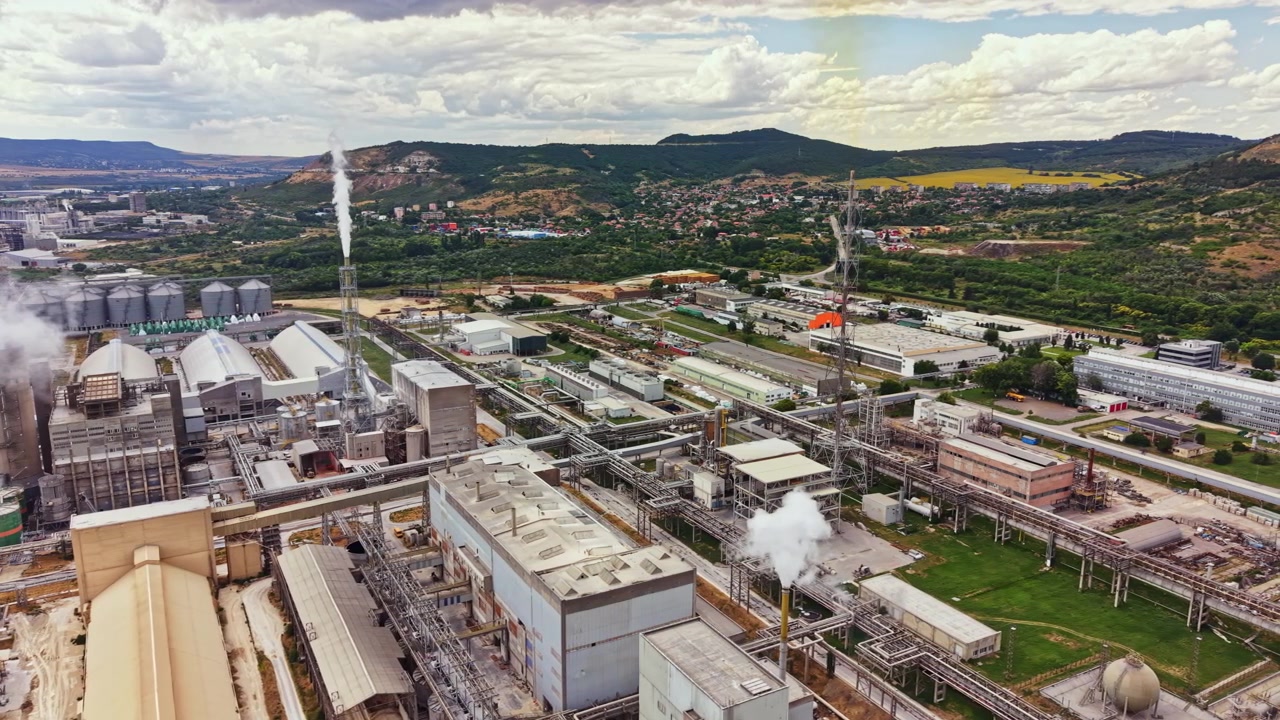 View of a large industrial complex with factories and warehouses located in a rural area under a cloudy sky near a mountain range during the daytime