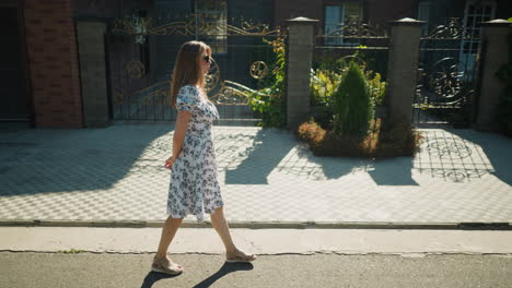 thoughtful woman in floral dress walking slowly with hands behind back along sunny suburban sidewalk, passing building with wrought iron gate and decorative paving under clear daylight