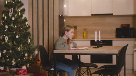 Side-View-Of-A-Girl-In-Green-Sweater-Writing-A-Letter-And-Thinking-Of-Wishes-Sitting-At-A-Table-In-A-Room-Decorated-With-A-Christmas-Tree