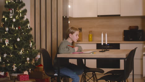 side view of a girl in green sweater writing a letter and thinking of wishes sitting at a table in a room decorated with a christmas tree