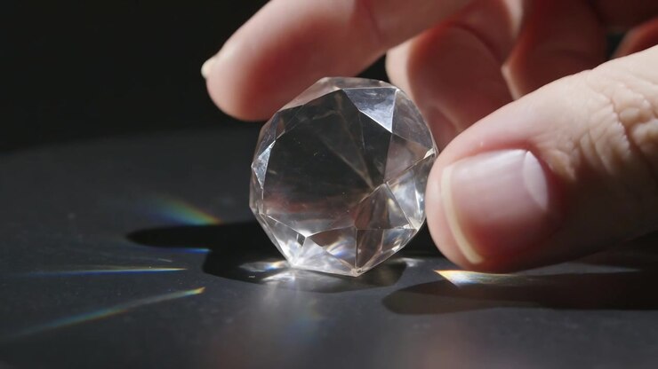 Una mano che si allunga inclina una sfera di cristallo sfaccettata su un tavolo scuro per modificare i fasci di luce prismatica.