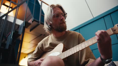 view from below: a blond man with long hair in glasses with a beard plays the electric guitar in a cozy room