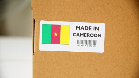 manos aplicando la etiqueta de la bandera hecha en camerún en una caja de cartón de envío con productos