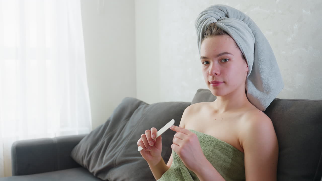 Woman sitting on couch with towel wrapped around hair and chest, holding nail file while looking relaxed and focused, enjoying quiet self-care moment in bright naturally lit home environment