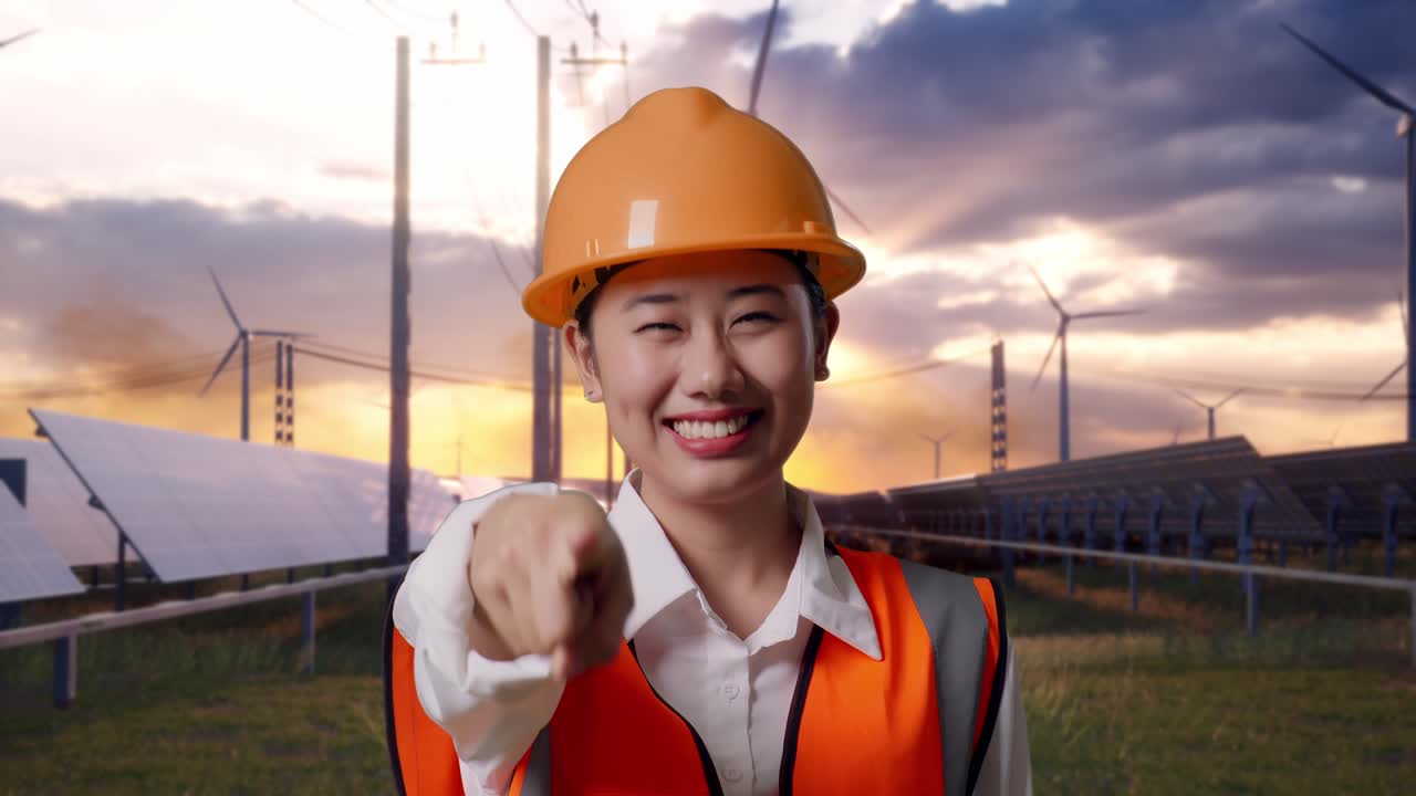 Close Up Of Asian Female Engineer With Safety Helmet Smiling And Touching Her Chest Then Pointing At You With Solar Panel and Wind Turbines