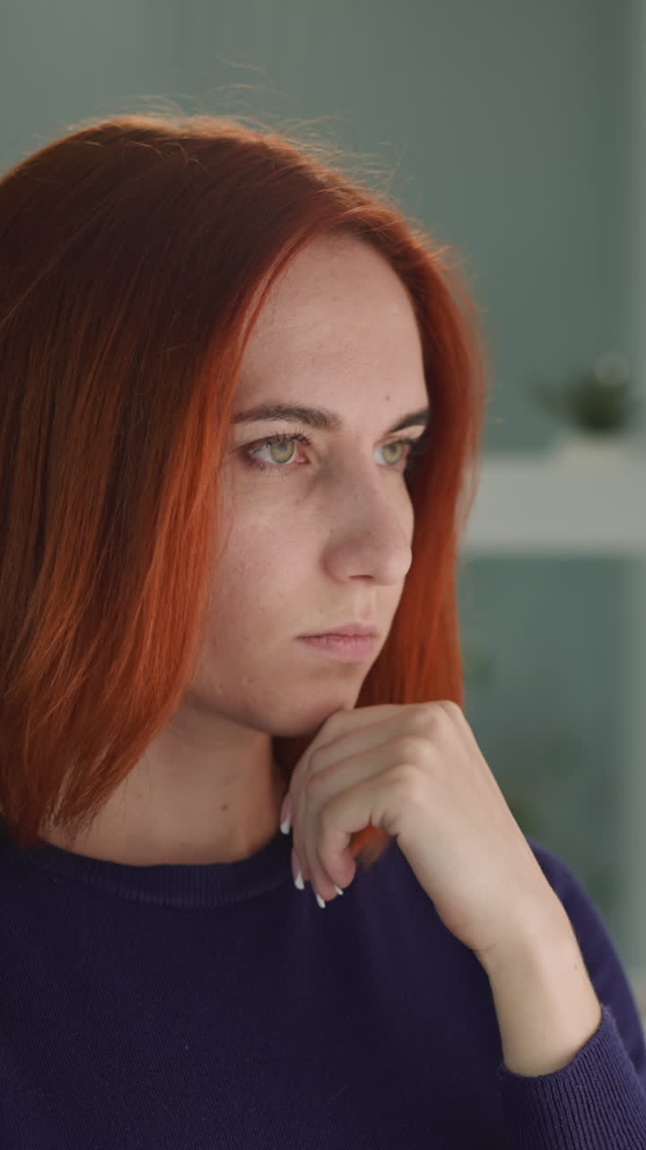 Pensive redhead woman looks out window touching chin with hand. Thoughtful lonely female thinks about meaning of life standing in bedroom closeup