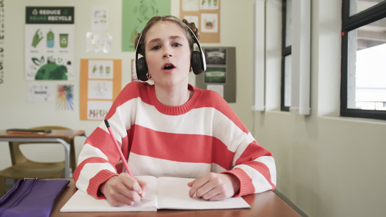 On video call, In school, young girl wearing headphones writing in notebook, smiling in classroom