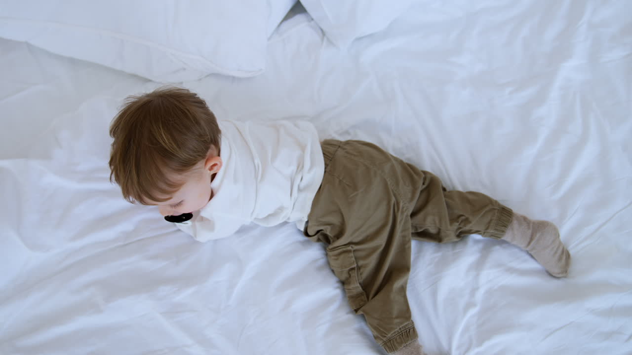 Small baby boy wearing white shirt and grey pants lies on a big bed. Toddler sucking a pacifier turns to his belly. Top view.