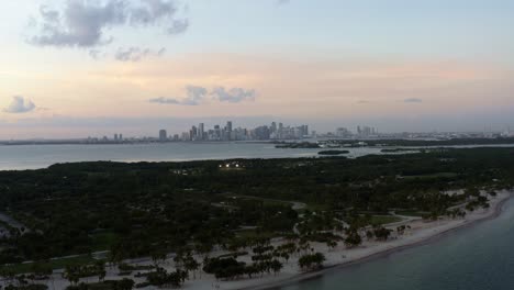 dolly out descending aerial drone shot of the tropical beach surrounded by palm trees on crandon park in key biscayne with the skyline of miami, florida in the distance on a sunny summer evening