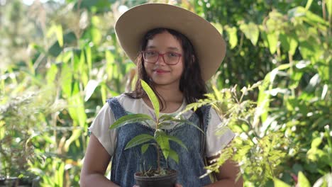 smiling girl wearing a hat, coverall and glasses, standing in the middle of a botanical garden with an avocado plant in her hands