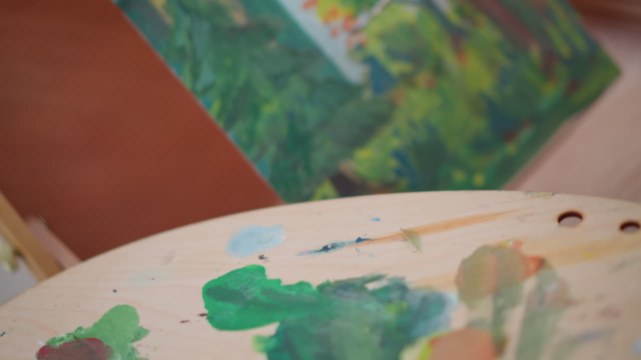 artista di strada che dipinge alberi con una palette su un'area di lavoro, che mescola i verdi su una palette di legno, mani che aggiungono la texture del fogliame a un paesaggio di cottage su un cavalletto, sfondo bokeh con luce solare calda, schizzo che si evolve in