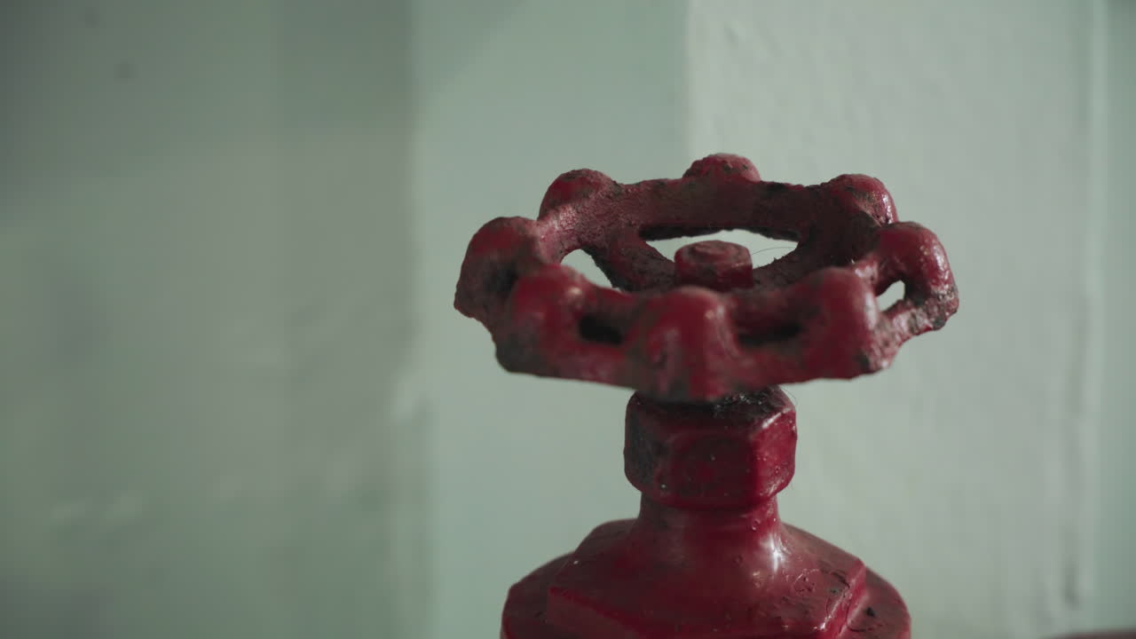 Maniglia a valvola rossa contro una parete chiara. Ruota in ghisa arrugginita con vernice scheggiata, superficie ruvida, sfondo verde tenue, elemento industriale fisso, contesto di manutenzione, atmosfera da ispezione idraulica
