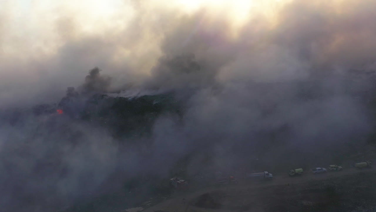 Aerial view around a group of Firefighters and firetrucks quelling a wildfire, in middle of dark smoke , sunny evening, in Los Angeles, California, USA - Orbit, drone shot