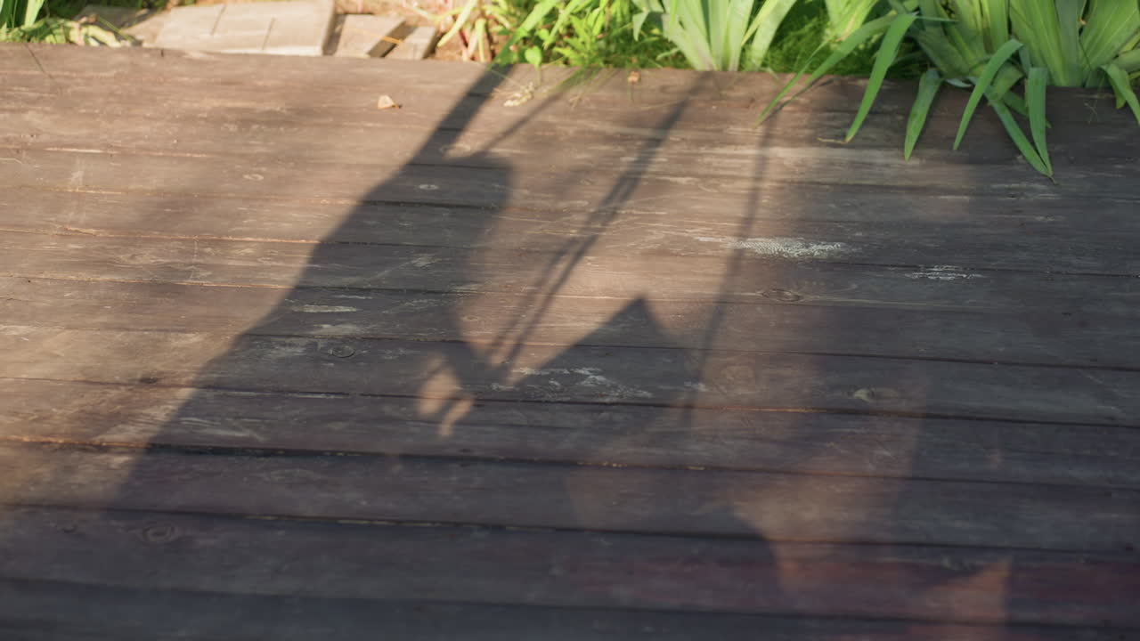 Player Swings Racket, Dynamic Scene Of Tennis Player Moving, Sunlit Wooden Deck Captures Playful Racket Swing, Elongated Shadows Depict Energetic Tennis Action On Sunny Textured Surface