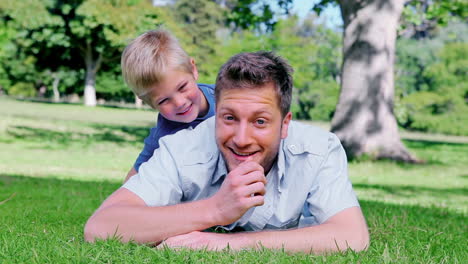 Father-looking-ahead-while-lying-on-the-grass-before-his-son-peeks-his-head-out-from-behind-him