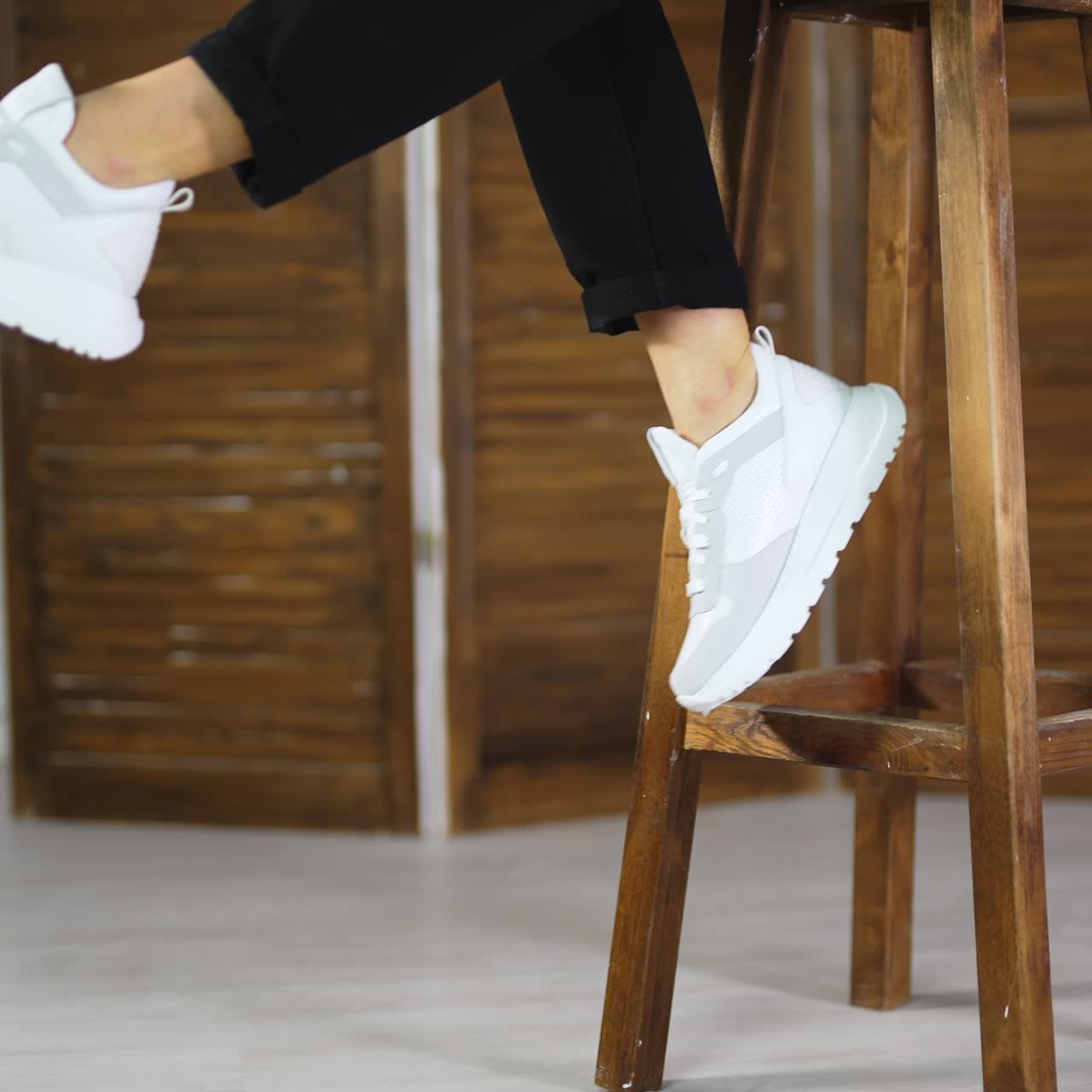 Model in black jeans comes to a wooden stool, sits on it and shows white new sneakers. Girl wave her legs to demonstrate fashionable footwear