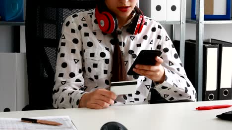 woman with red headphones on shoulders holding credit card and using smartphone