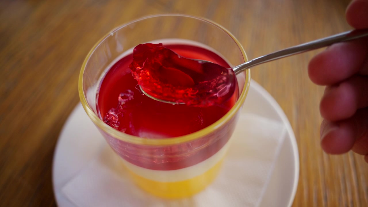 Primo piano di un cucchiaio che solleva un pezzo di gelatina di frutta rosso vivo da una coppetta da dessert di plastica trasparente.
