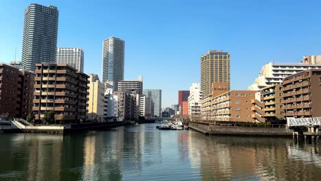 sunlit cityscape with modern buildings along a calm river, clear blue sky, urban waterfront scene