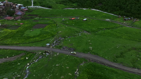 Vista-Aérea-De-La-Conducción-De-Automóviles-En-Un-Camino-Sin-Pavimentar-En-Medio-De-Un-Prado-Verde-Hacia-El-Pueblo-De-Ushguli-En-Georgia