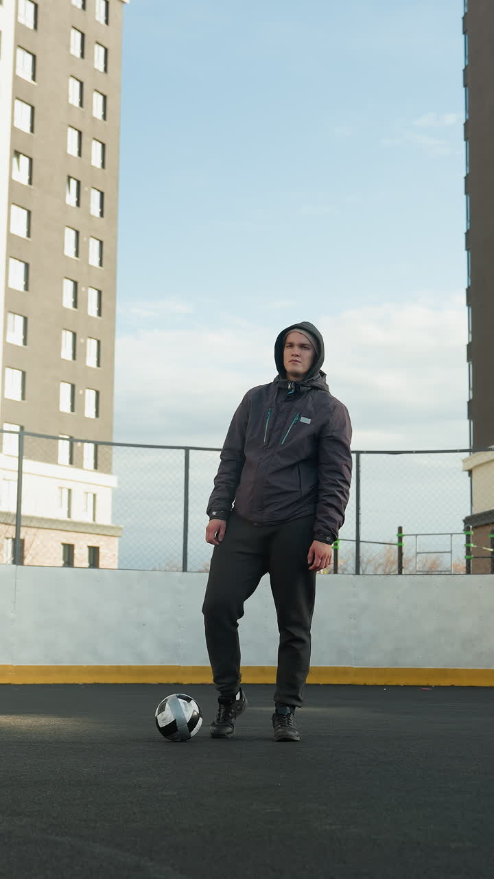 Sportsman in hoodie and athletic wear practicing ball control, standing on sport arena field with goalpost behind, urban background highlights tall modern office buildings