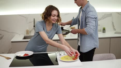 pareja alegre en la cocina blanca y brillante. prepararon tortilla en la sartén, la novia está sirviendo el plato, agregando tomates. emocionado y hambriento. cámara lenta