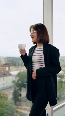 modern young lady in black jacket and jeans stands at the window. woman drinks coffee and enjoys city view. vertical video.